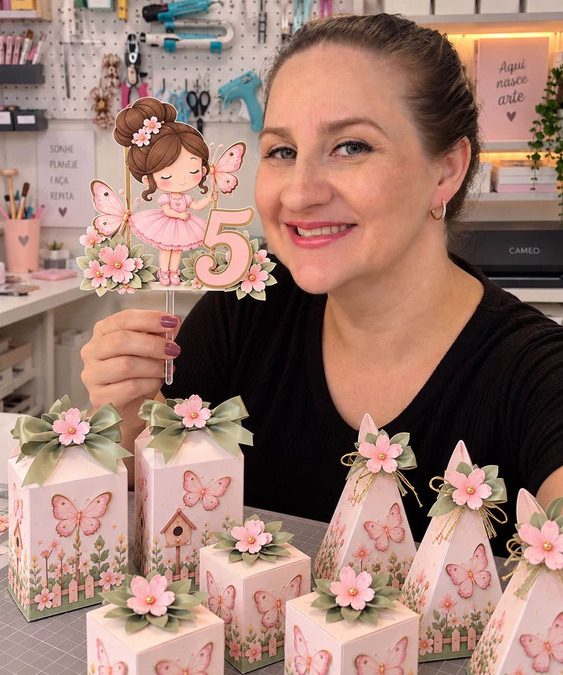 Rafa Calcic segurando topo de bolo personalizado no ateliê, cercada de caixinhas decoradas
